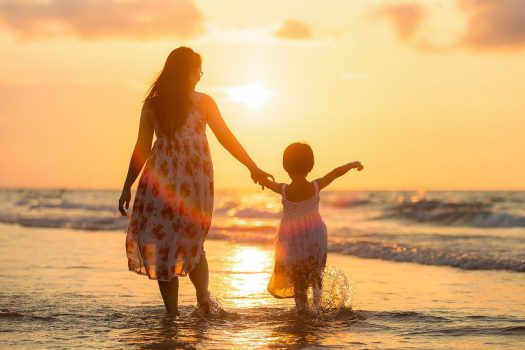 Ibu menggandeng anak perempuan di pinggir pantai dengan background matahari terbenam
